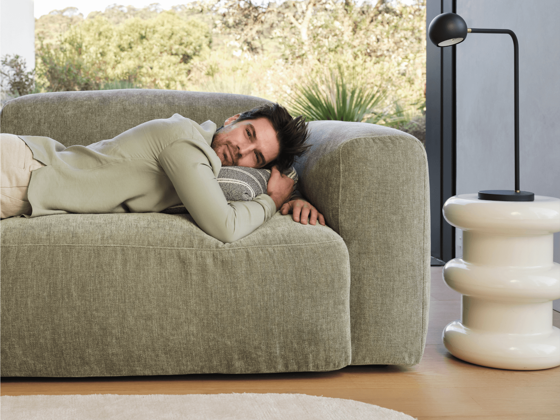 Modern sage green linen sofa with plush cushions and a minimalist design in a sunlit room, complemented by a white glossy round side table.