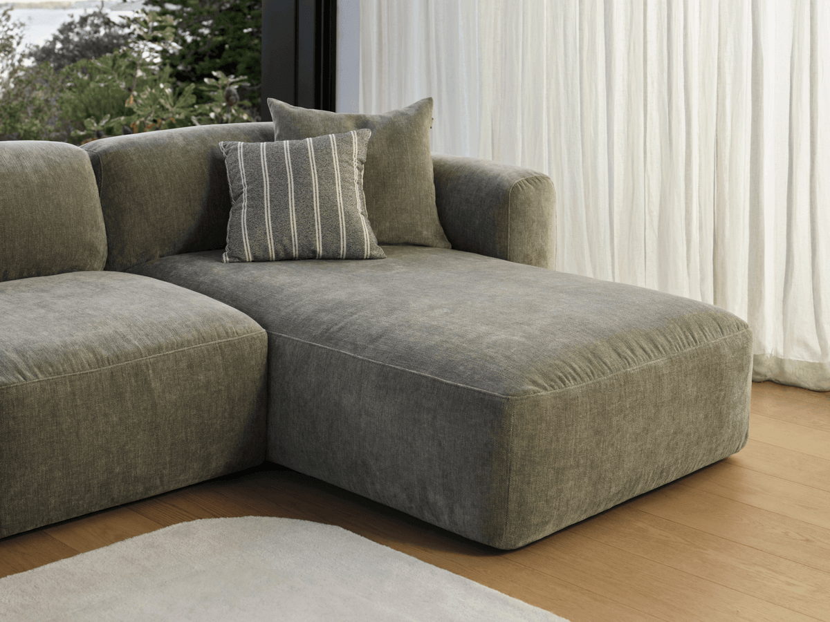 Luxurious grey linen chaise lounge with plush cushions, set on polished wooden floors, adjacent to sheer white curtains in a modern living space.