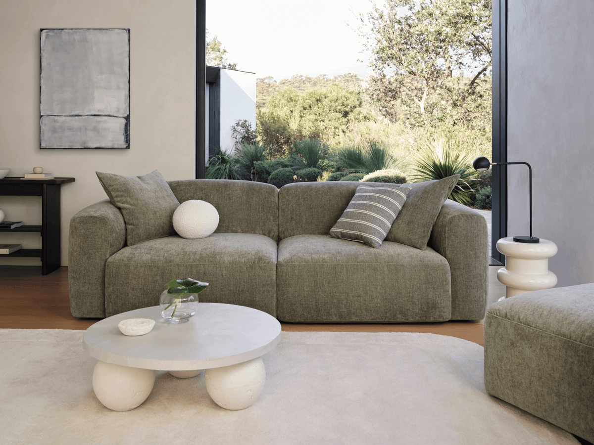 Modern olive green sofa with plush cushions and soft upholstery, set in an elegant living room. Features a minimalist coffee table and scenic garden view.