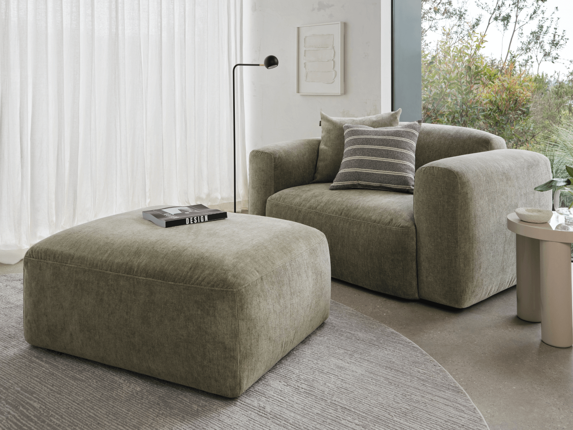 Curved olive green lounge chair and matching ottoman in plush fabric. Set on a grey rug, near sheer curtains and a modern floor lamp.