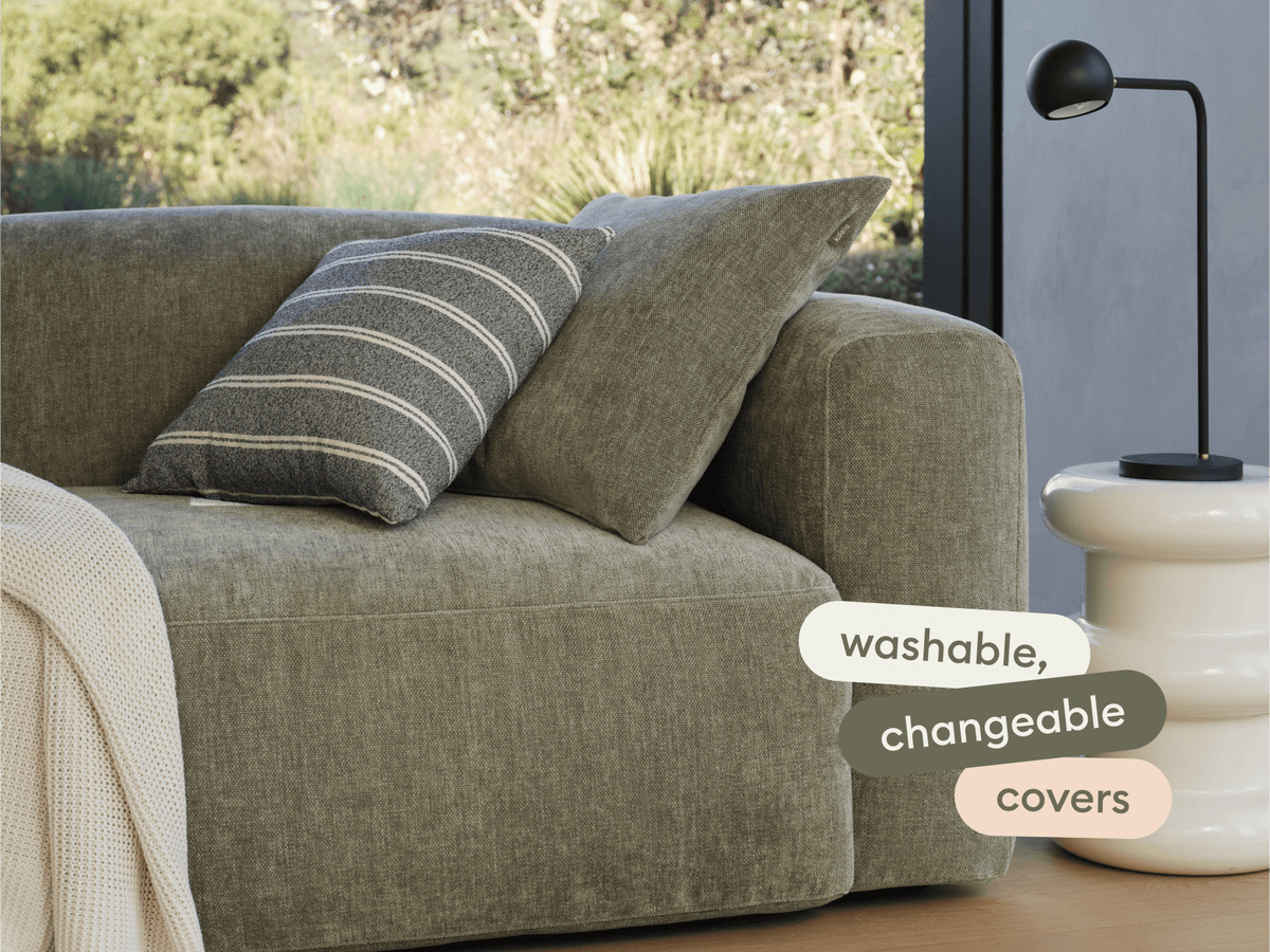 Luxurious olive green sofa with washable, changeable fabric covers, set in a modern living space. Features oversized cushions and a sleek white side table.