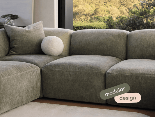 Luxurious modular sofa in soft grey fabric, featuring plush cushions and sleek lines. Positioned in a modern living room with large windows and garden views.