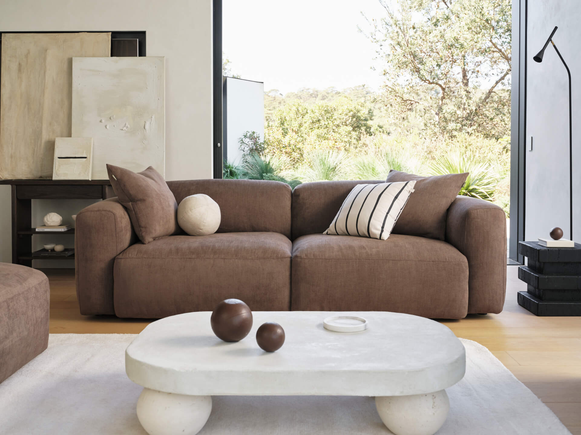 Luxurious brown fabric sofa with plush cushions, paired with a smooth white concrete coffee table. Set in a minimalist living room with floor-to-ceiling windows.