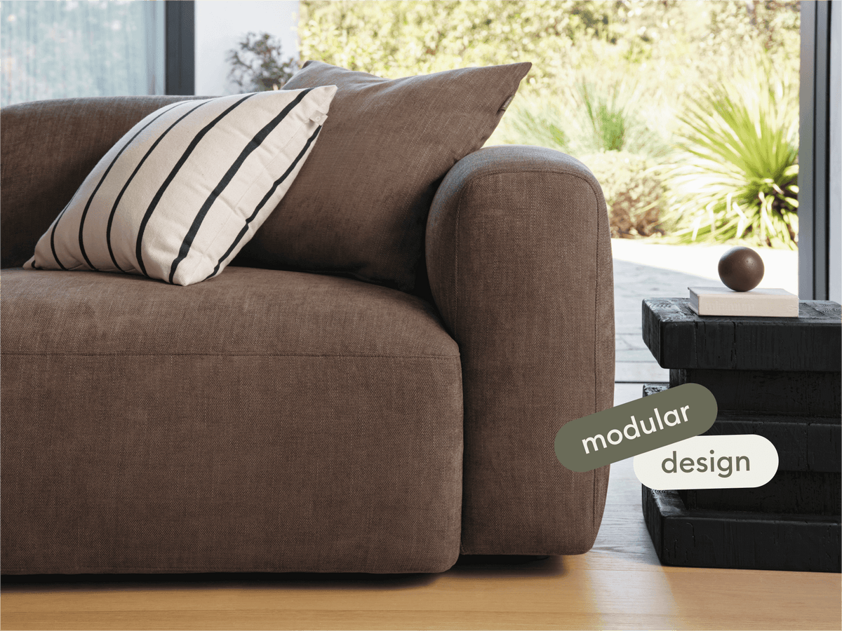 Luxurious brown fabric sofa with modular design, plush cushions, and a minimalist style. Situated in a modern living room with a natural outdoor view.