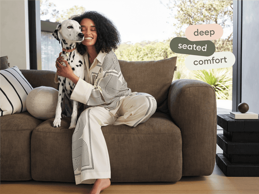 Luxurious deep-seated sofa in rich brown fabric, featuring plush cushions and curved armrests. Set in a bright, modern living room with natural light.