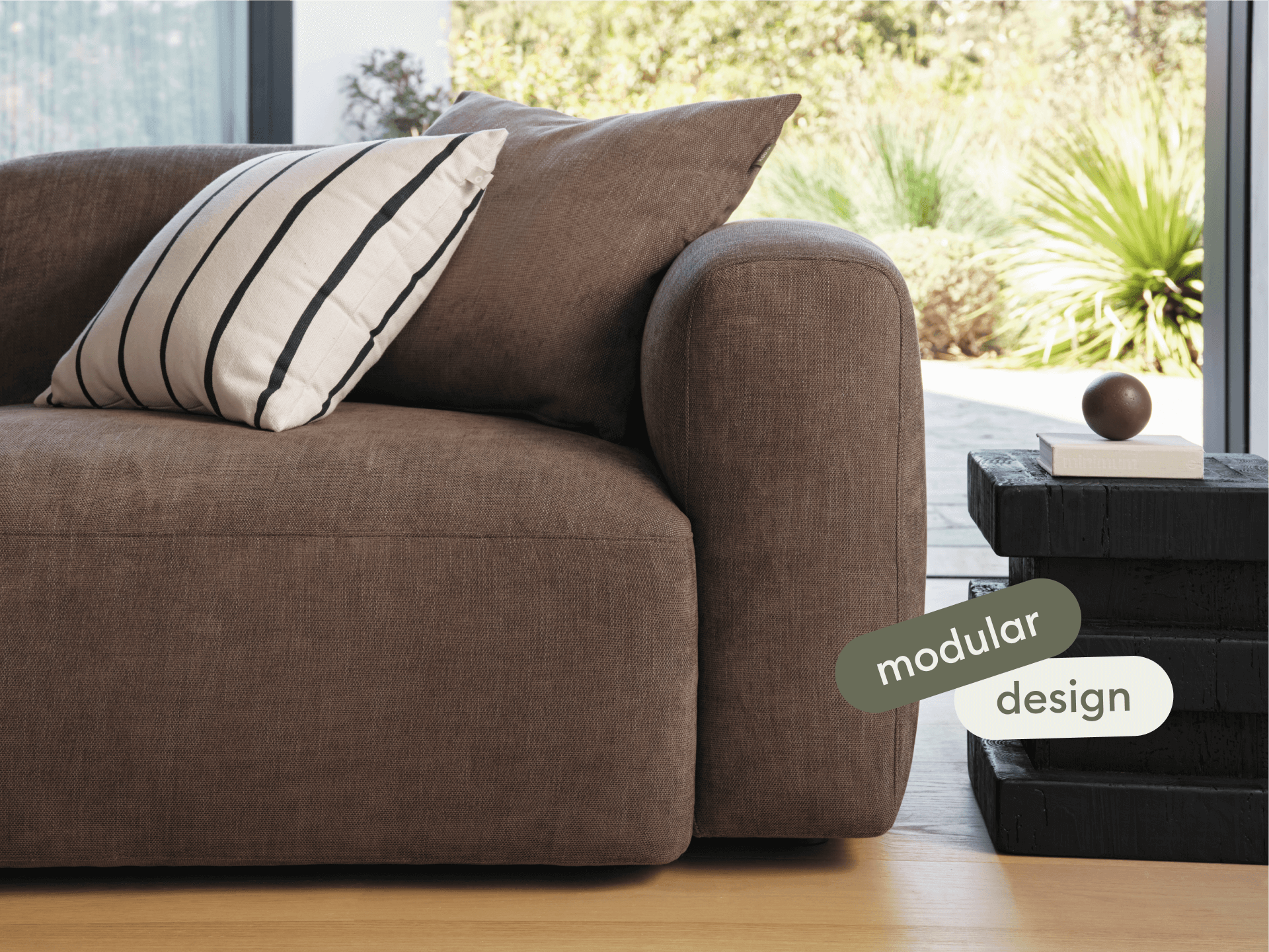 Luxurious brown fabric modular sofa with plush cushions and rounded armrests, set on light wooden flooring. Paired with a chic black side table.
