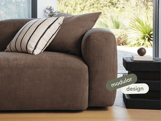 Luxurious brown fabric modular sofa with plush cushions and rounded armrests, set on light wooden flooring. Paired with a chic black side table.