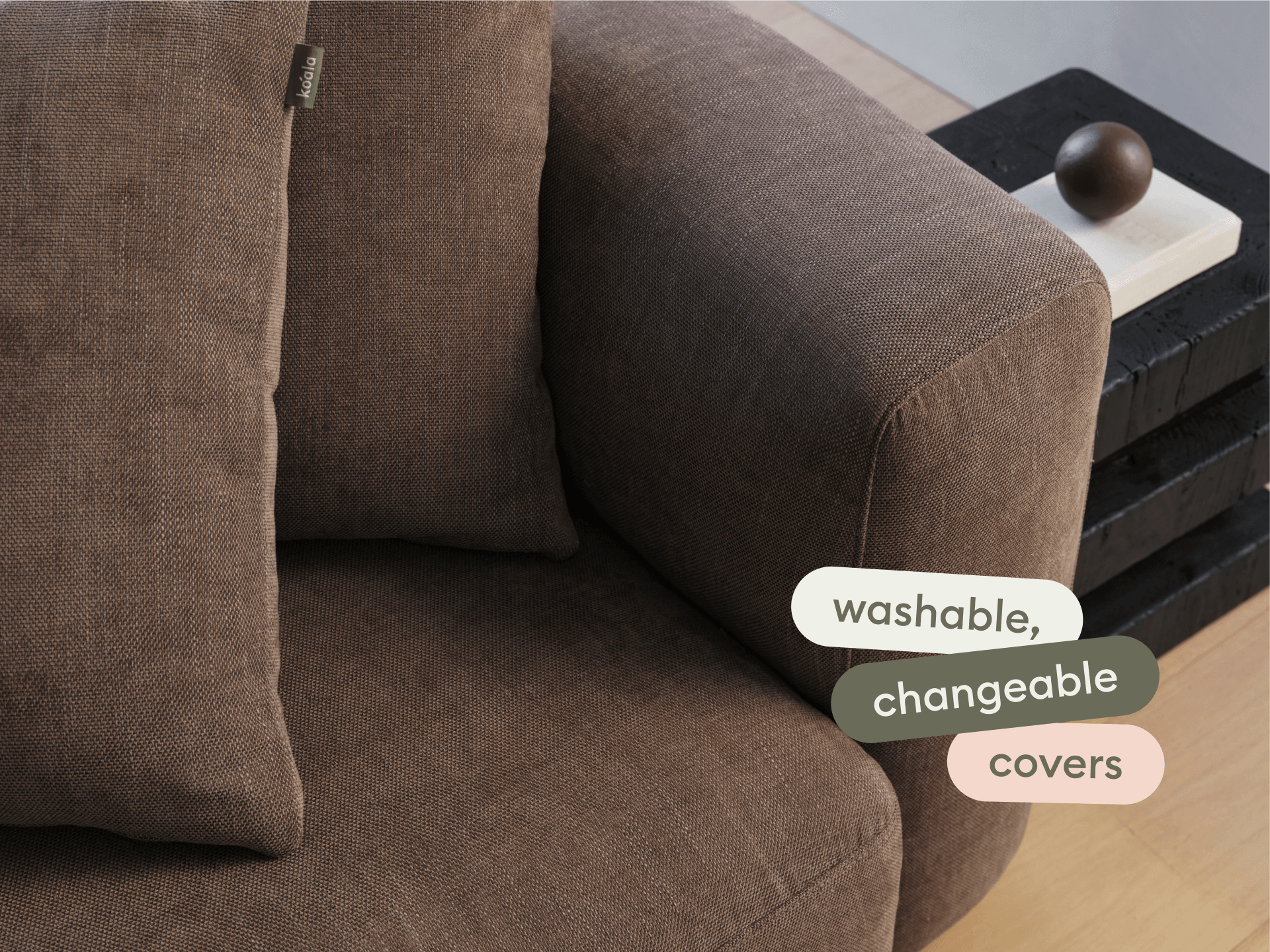Elegant brown fabric sofa with washable, changeable covers. Features plush cushions and wide armrests, set in a minimalist living space with wooden flooring.