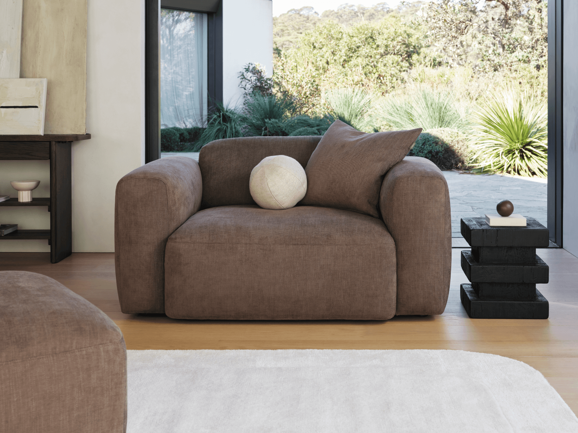 Luxurious brown linen armchair with plush cushions and wide armrests, set in a bright living room with floor-to-ceiling windows and a serene garden view.