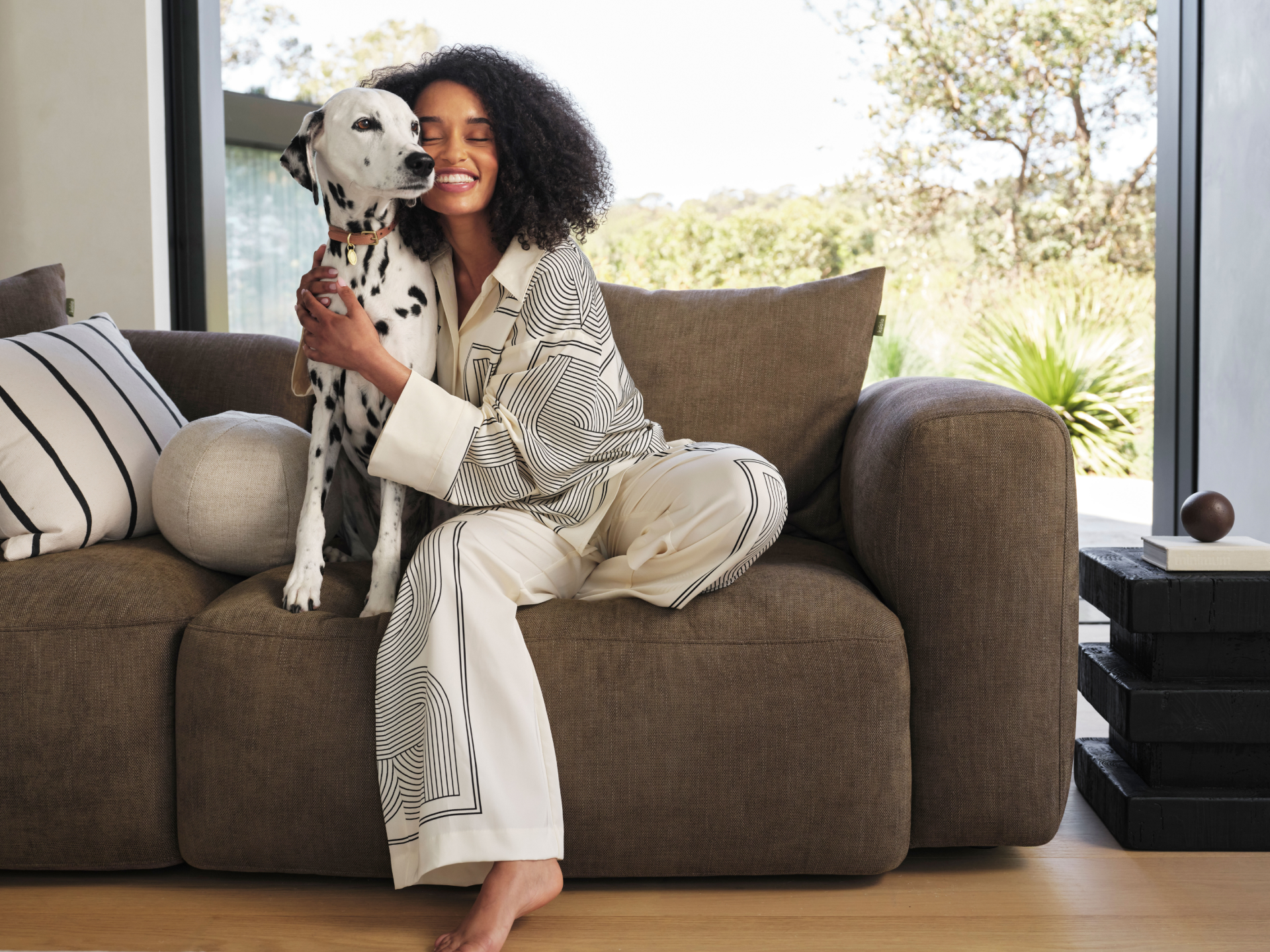 Luxurious brown fabric sofa with plush cushions, set in a bright living room with oak flooring and glass doors offering a serene outdoor view.