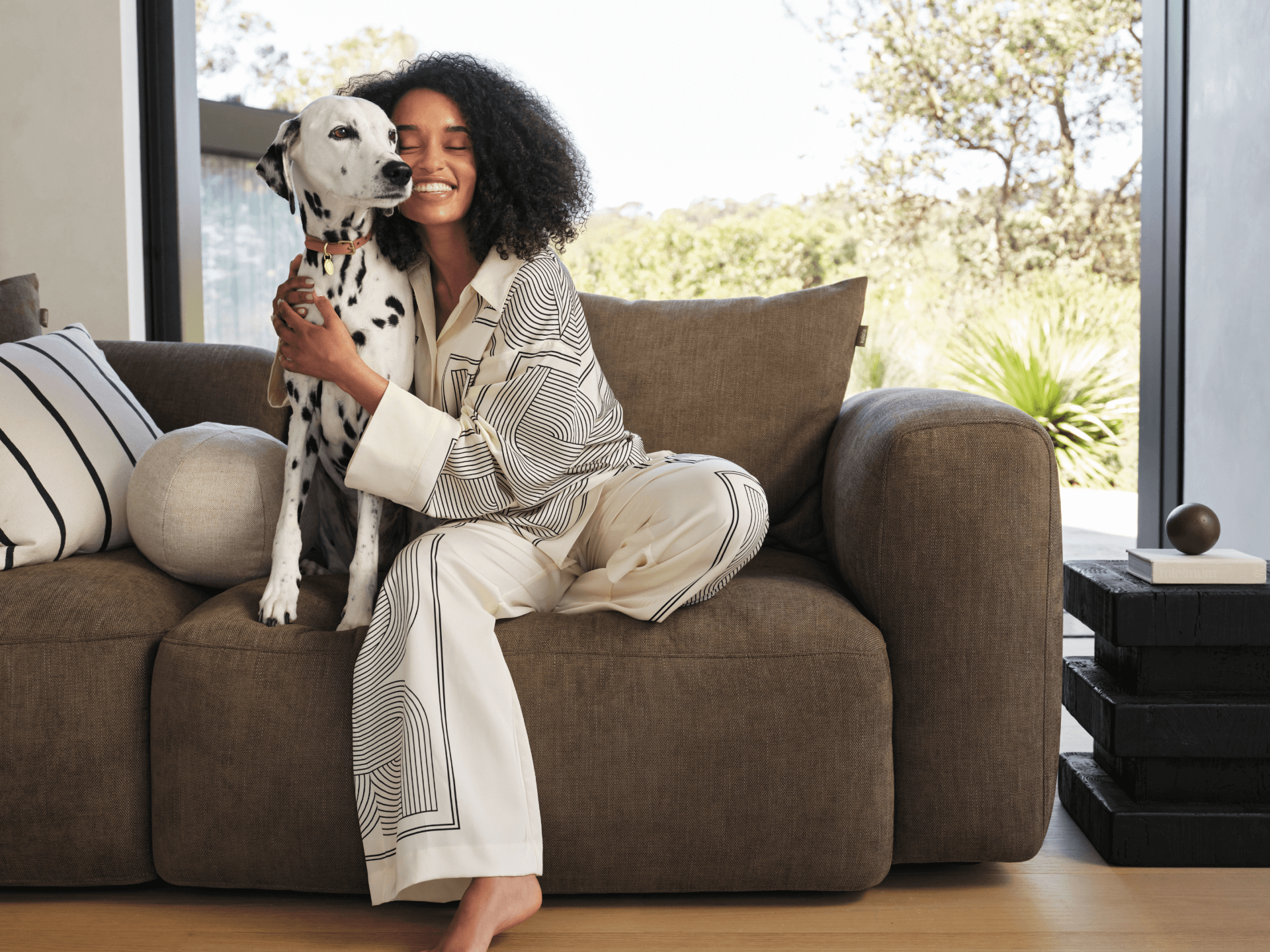 Luxurious brown fabric sofa with plush cushions, set in a sunlit living area featuring large glass doors. Neutral tones create a serene, modern ambiance.