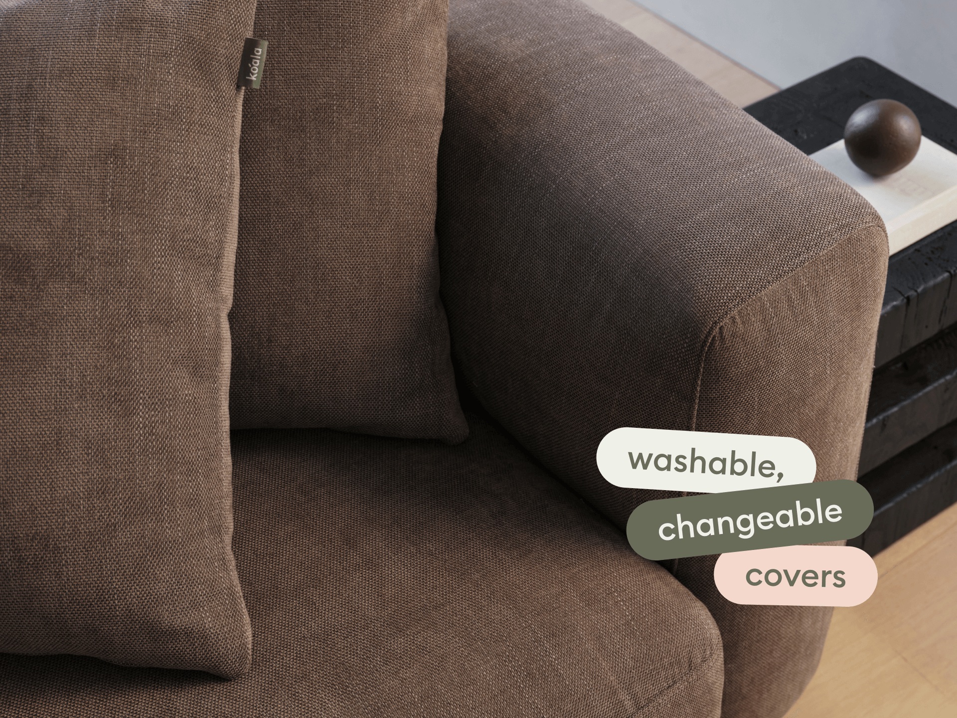 Modern sofa in rich brown fabric with washable, changeable covers. Features a plush cushion and sleek armrest. Set on light wood flooring.