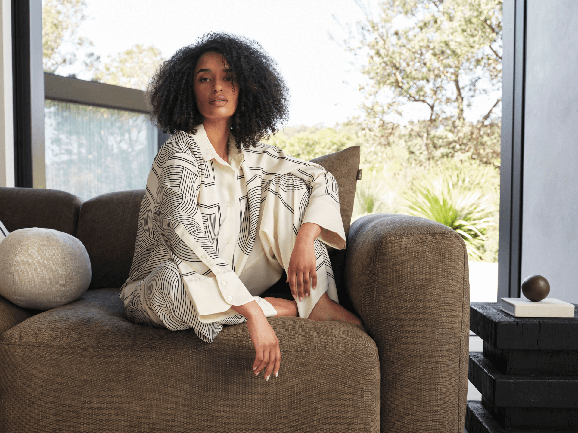 Luxurious brown linen sofa with plush cushions, featuring a modern design. Positioned in a sunlit, open-plan living area with expansive glass windows.