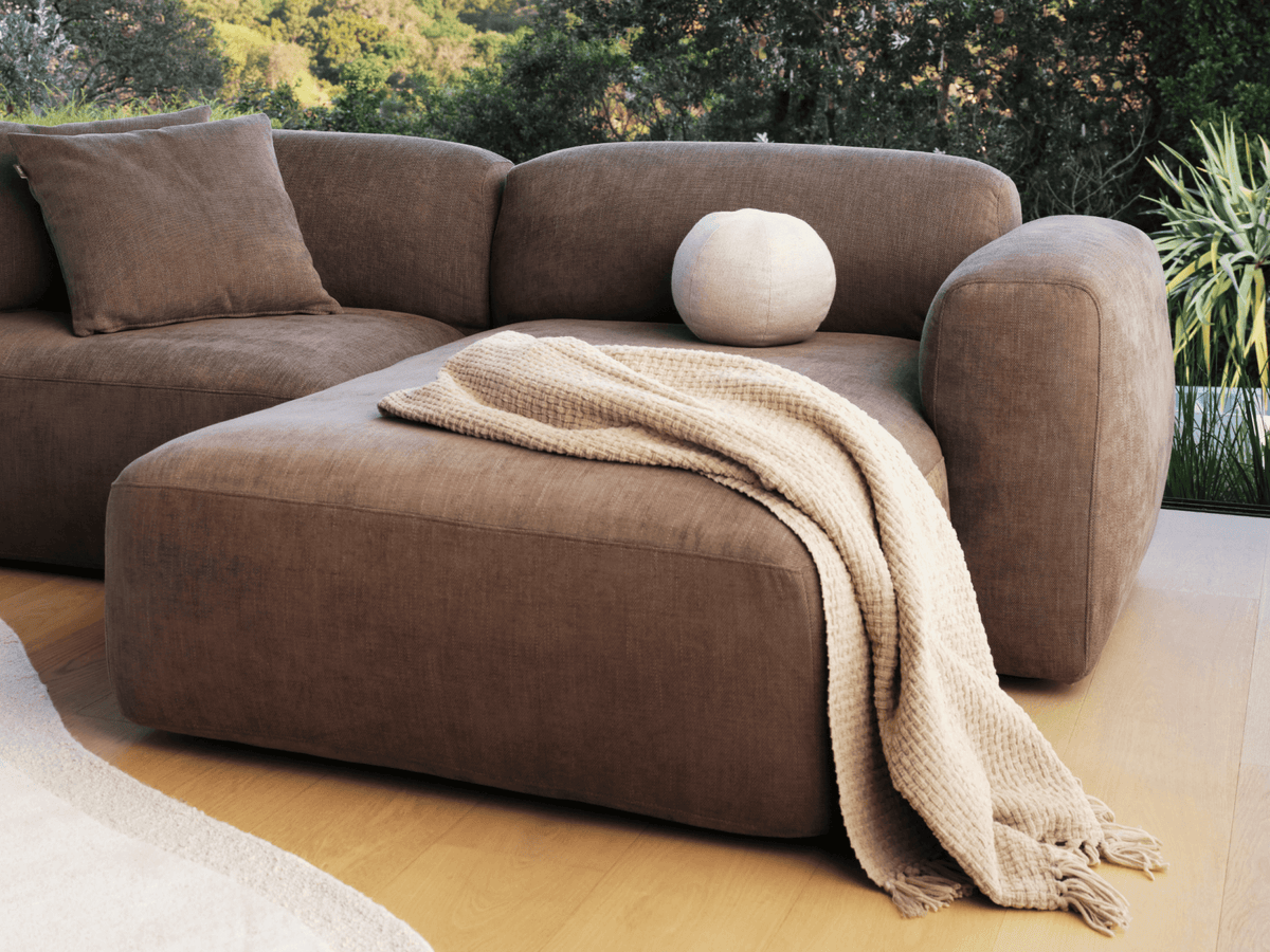 Luxurious brown fabric sectional sofa with a plush, modern design. Features a chaise and soft curves, set in a light-filled contemporary living space.