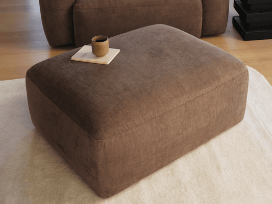 Luxurious brown fabric ottoman on a plush cream rug, featuring a sleek rectangular design and a smooth, soft-touch finish, perfect for contemporary interiors.