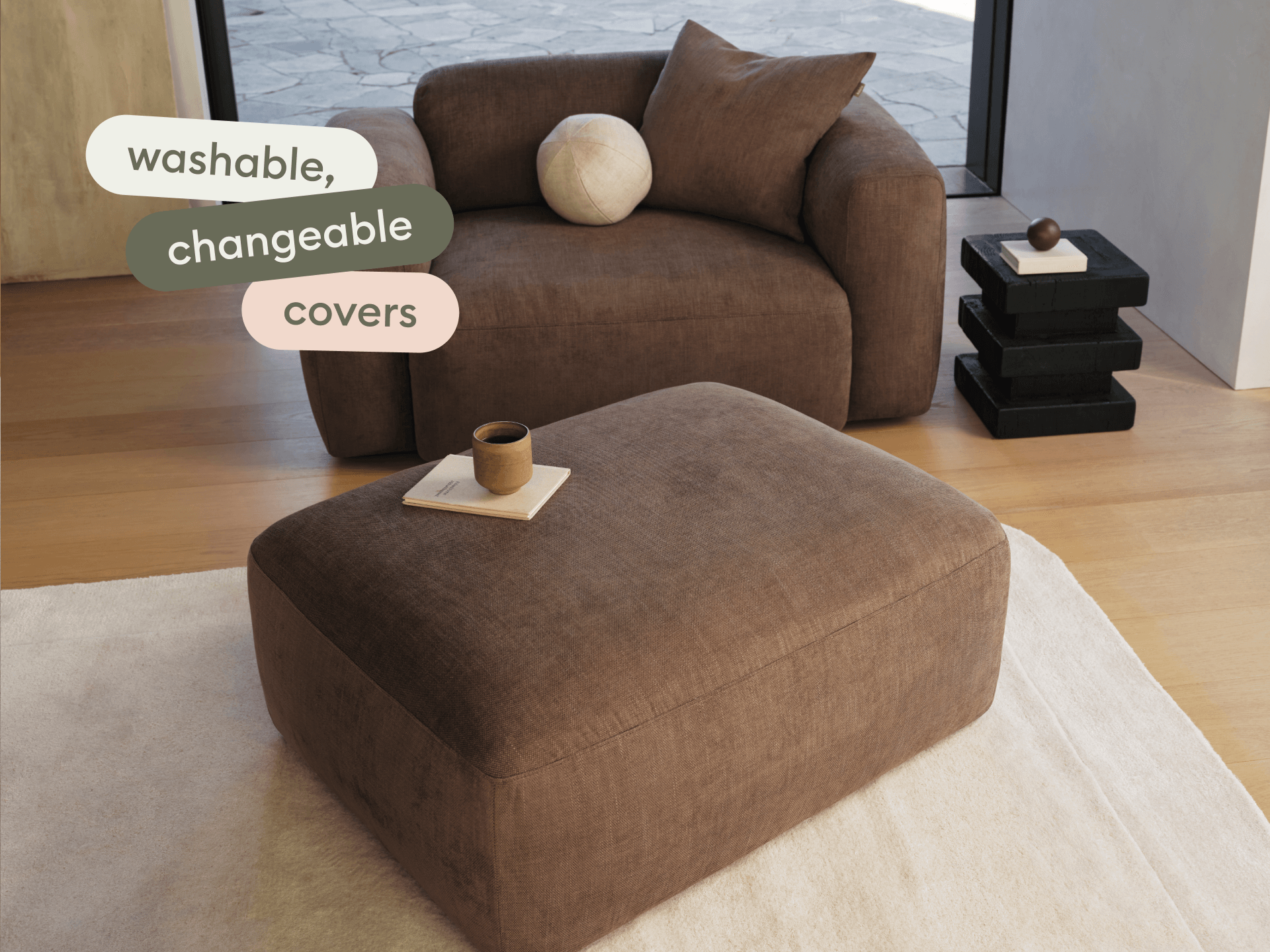 Plush brown fabric sofa with matching ottoman, featuring washable covers. Set on light timber flooring beside a minimalist black side table.