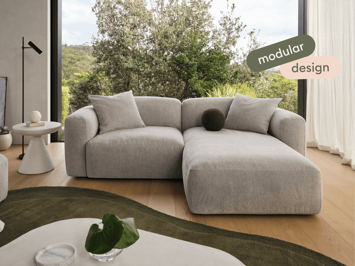 Luxurious grey modular sofa with plush fabric upholstery, featuring a versatile chaise lounge. Set in a light, airy living room with nature views.