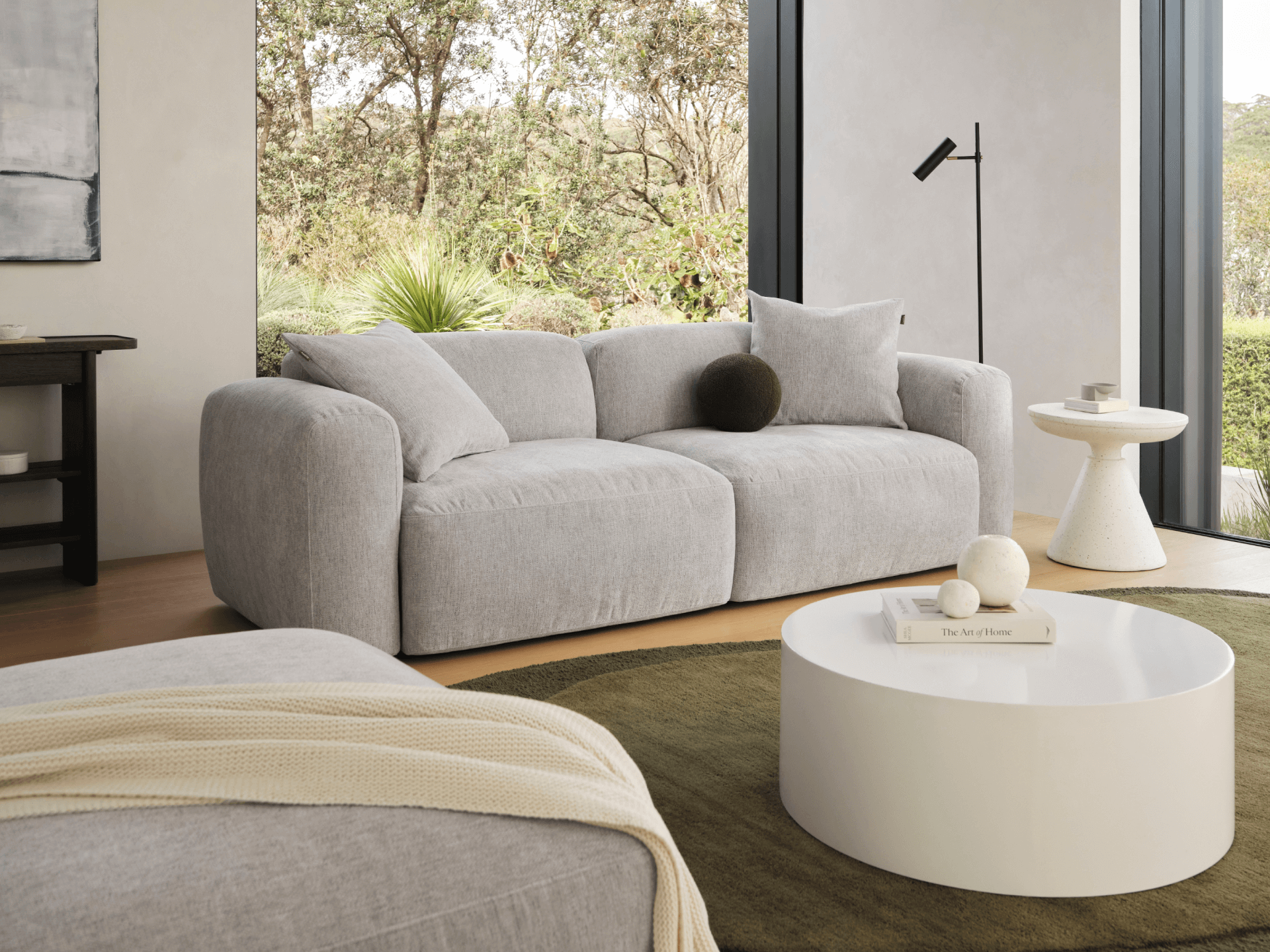 Grey upholstered sofa with rounded arms and plush cushions in a light-filled modern living room, complemented by a white round coffee table and olive-green rug.