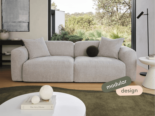Modern grey modular sofa with plush fabric upholstery and cushioned armrests. Set in a luxurious living room featuring sleek décor and lush garden view.