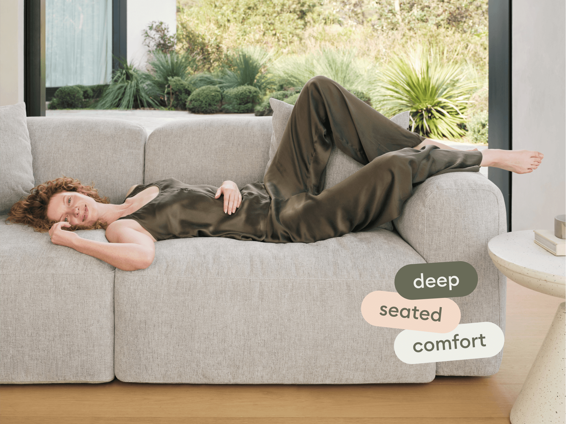 Modern grey fabric sofa with plush, deep seating and soft cushions. Set in a light-filled room with floor-to-ceiling windows, showcasing lush greenery outside.