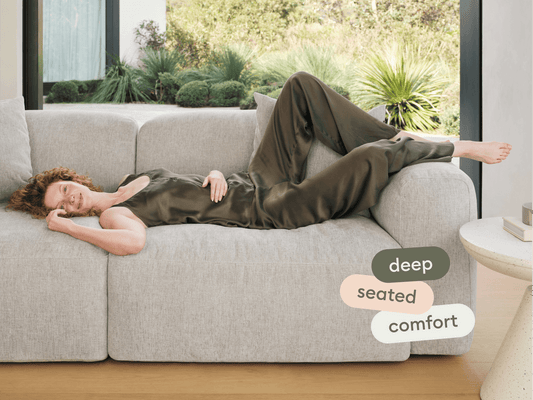 Modern grey fabric sofa with plush, deep seating and soft cushions. Set in a light-filled room with floor-to-ceiling windows, showcasing lush greenery outside.
