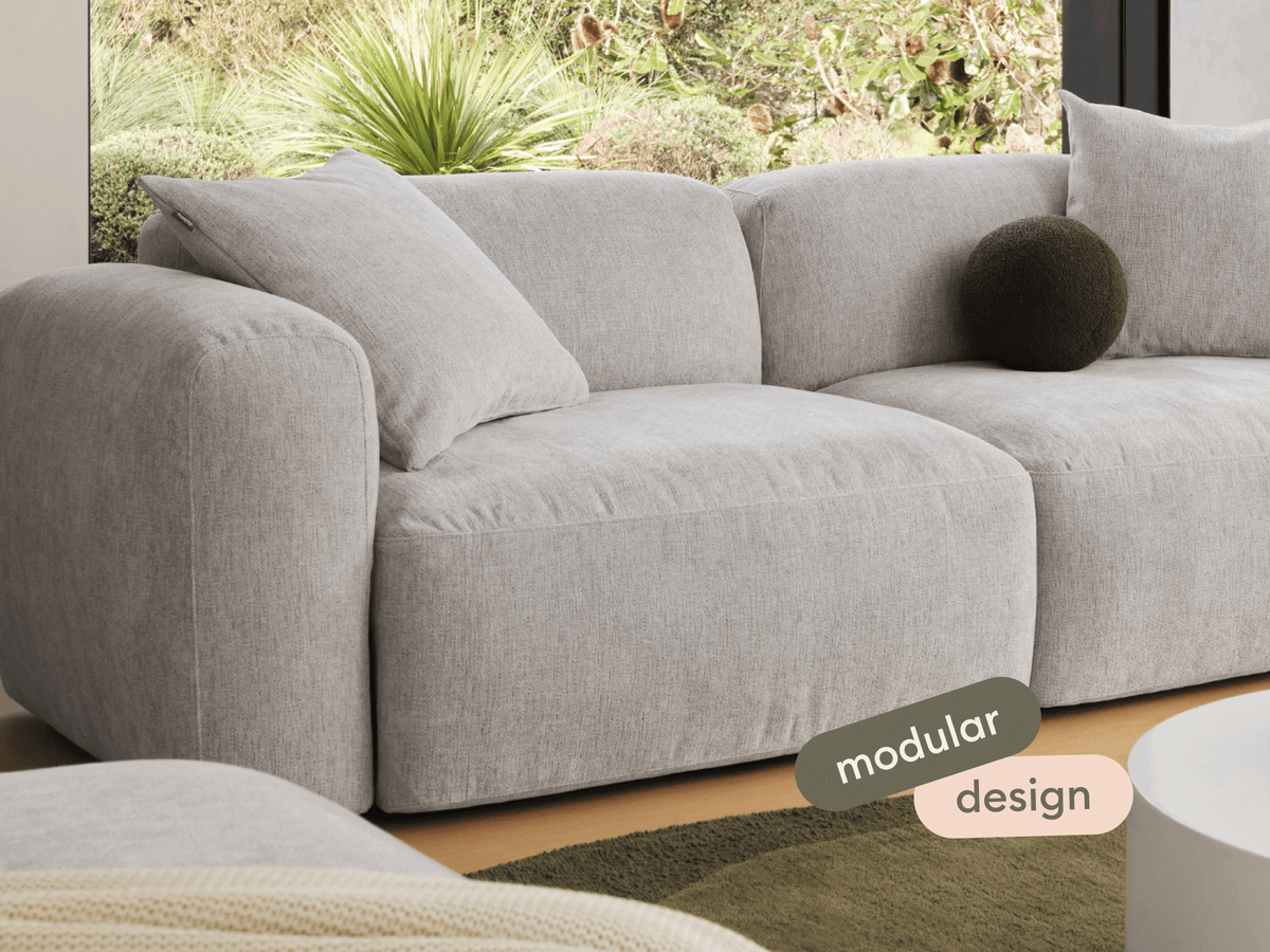 Sophisticated grey fabric modular sofa with plush cushions and rounded arms, set in a sunlit living area with modern décor and lush garden views.