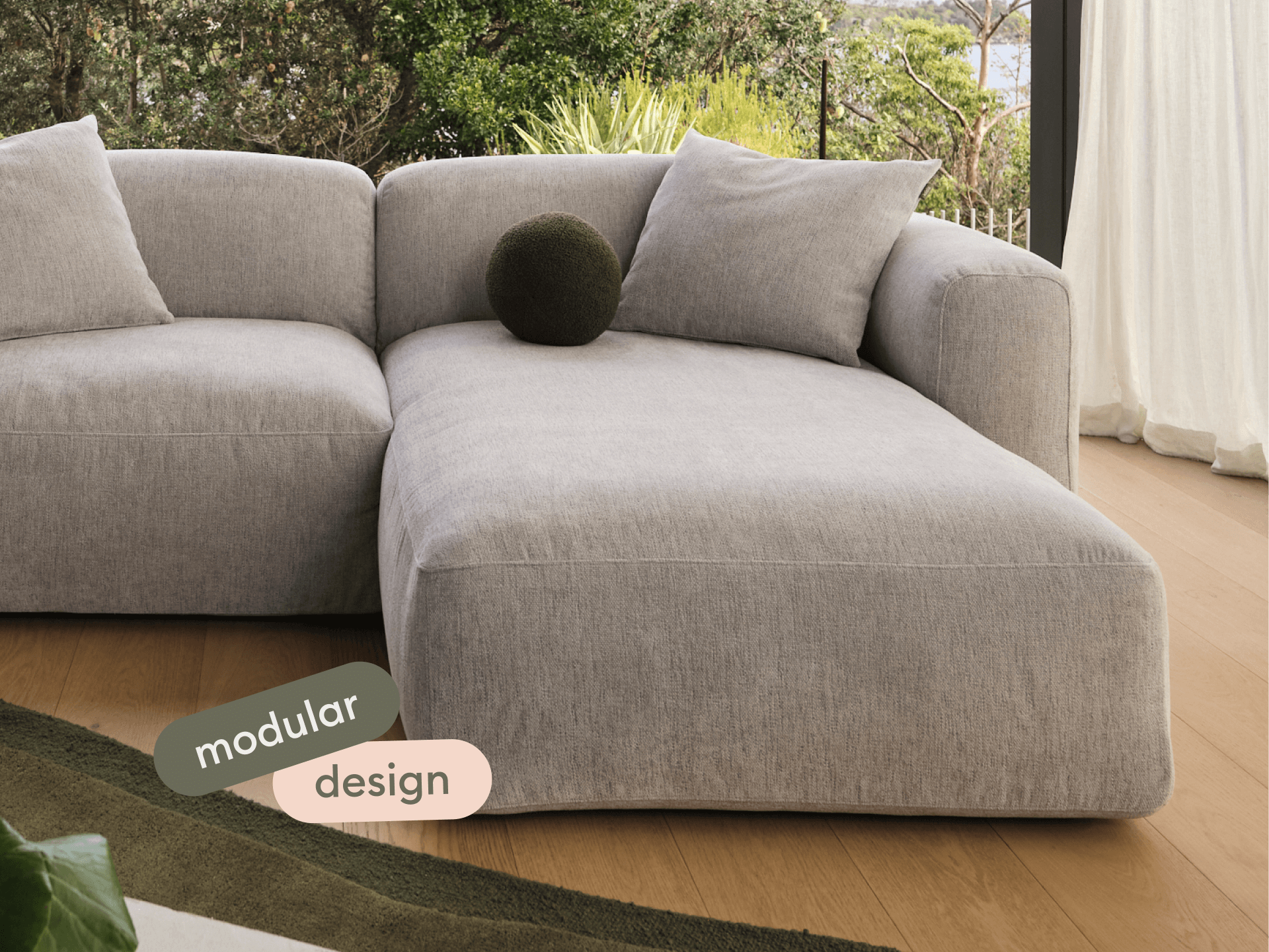 Luxurious modular sofa in soft grey fabric, featuring a sleek chaise section. Set on elegant timber flooring with lush greenery in the background.
