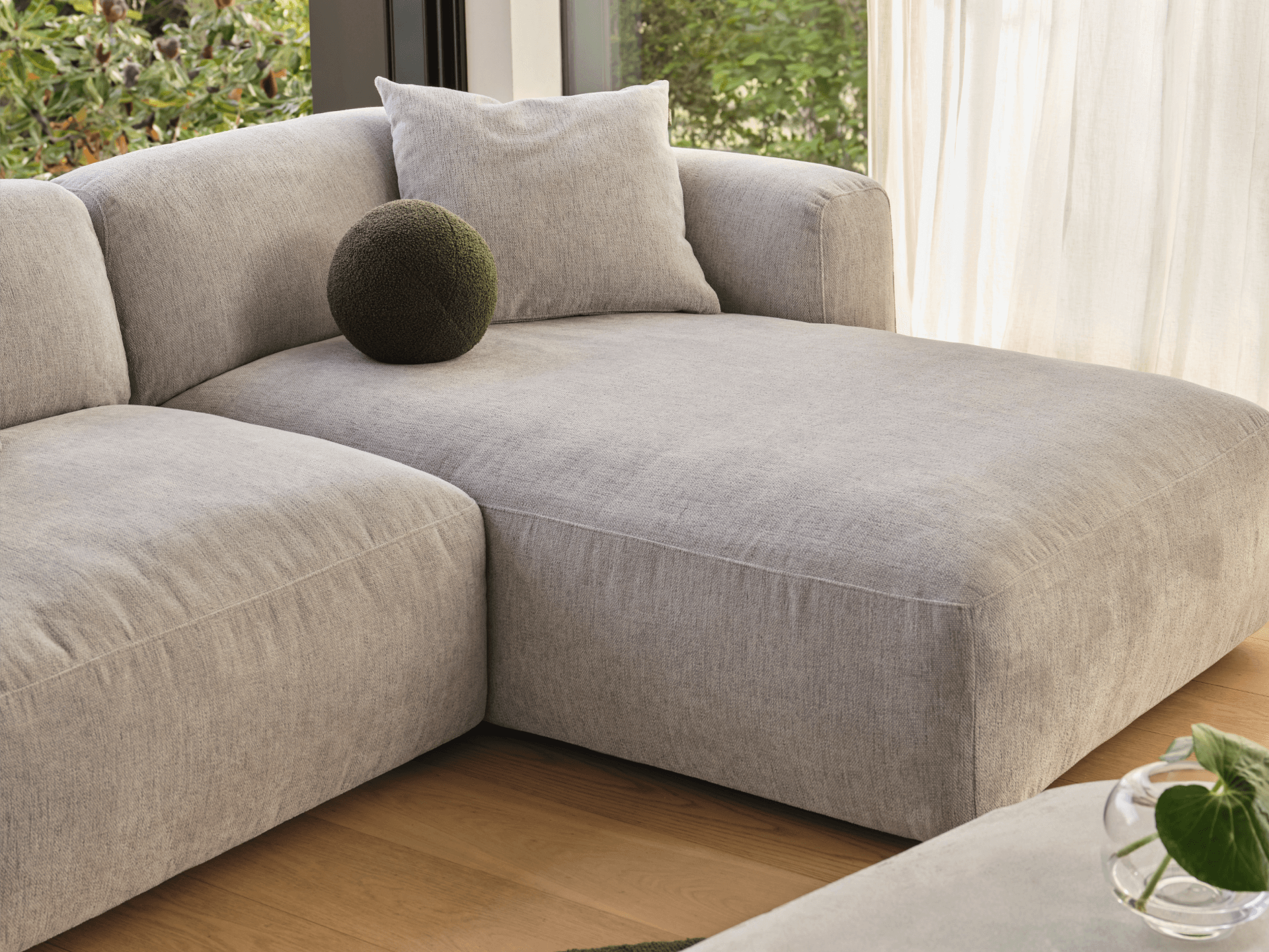 Luxurious grey linen sectional sofa with plush cushions, streamlined design, and clean lines. Set in a sunlit room with wooden flooring.