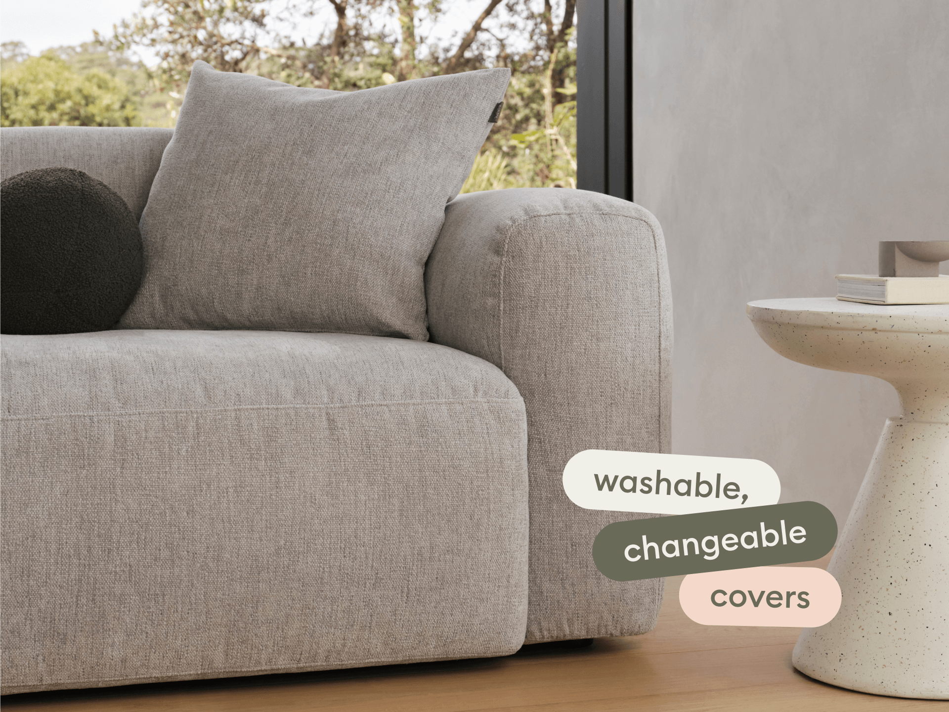 Luxurious grey fabric sofa with plush, boxy cushions and washable, changeable covers. Set in a minimalist room with light wood flooring and a stone side table.