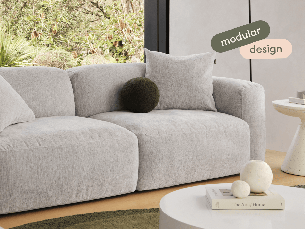 Modern grey modular sofa in soft fabric, featuring plush cushions and rounded corners. Set in a light, airy room with garden views, textured accents, and a chic ambience.