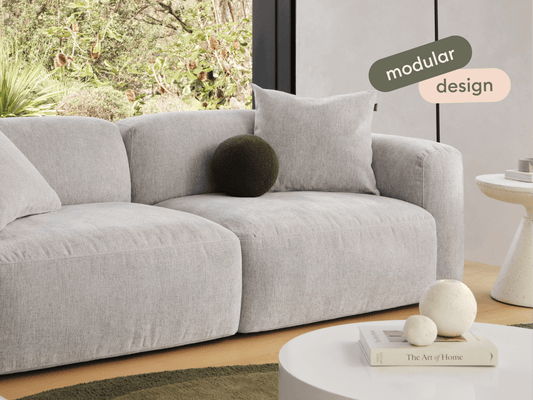 Modern grey modular sofa in soft fabric, featuring plush cushions and rounded corners. Set in a light, airy room with garden views, textured accents, and a chic ambience.