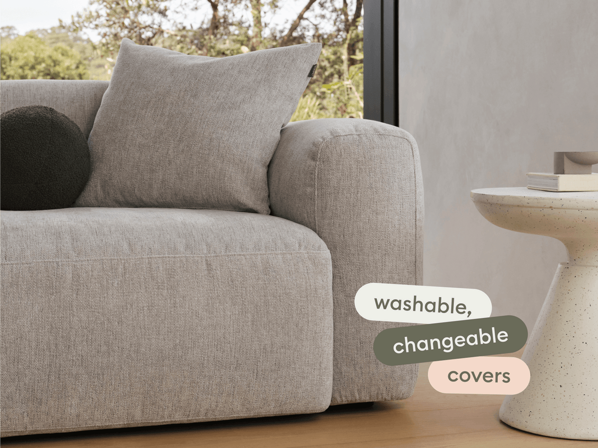 Modern grey fabric sofa with plush, washable covers and substantial armrest, paired with a minimalist stone side table. Set against a sunlit interior.
