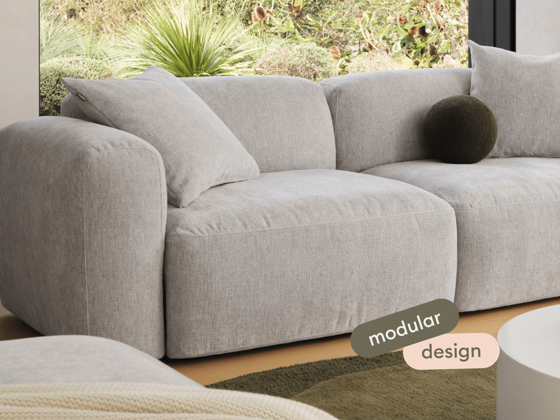 Luxurious modular sofa in soft grey fabric, featuring plush cushions and rounded armrests. Set against a minimalist backdrop with lush greenery outside.