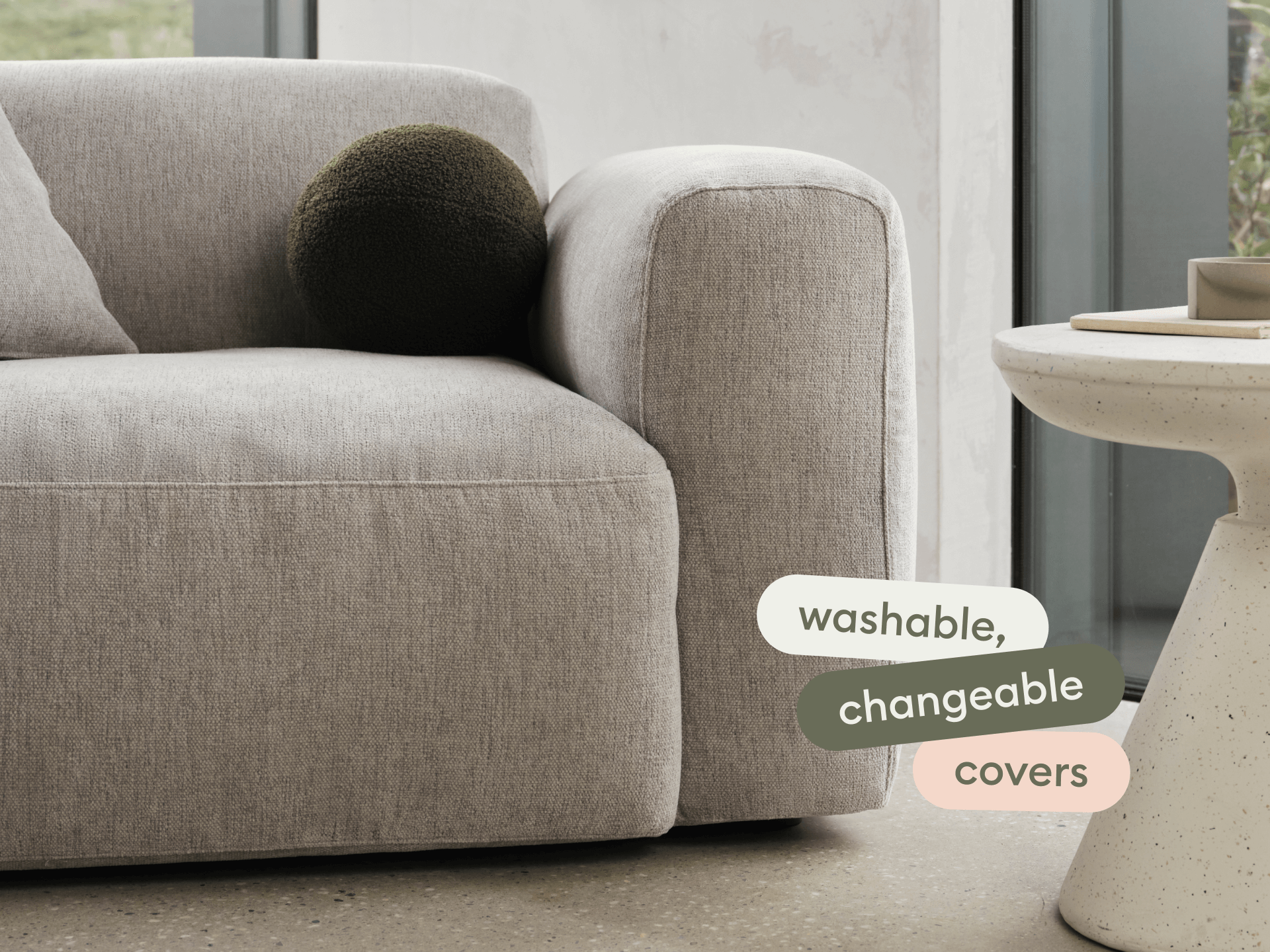 Contemporary grey fabric sofa with plump cushioning and washable, changeable covers. Set beside a modern, round white side table in a minimalist living space.
