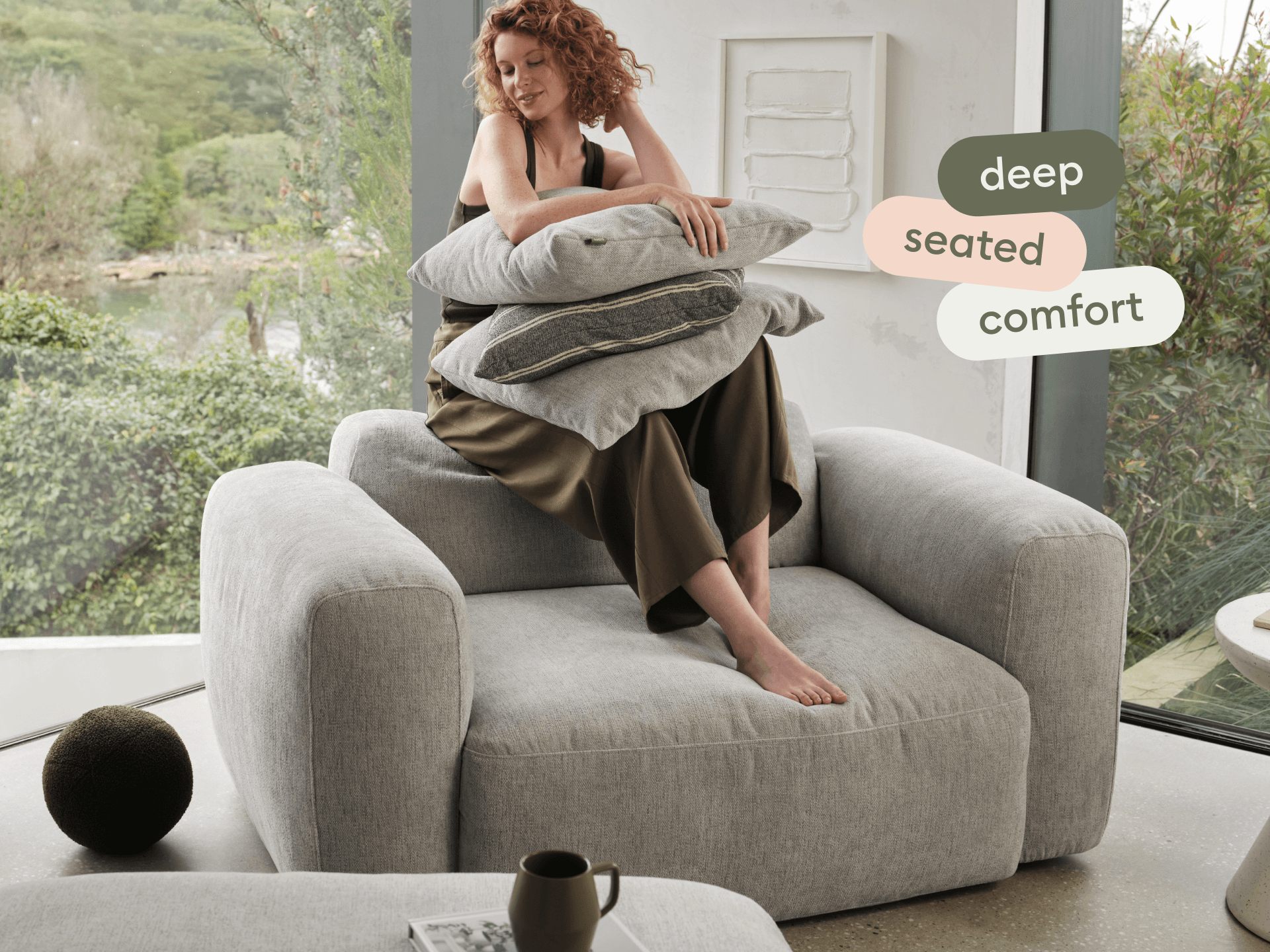 Luxurious grey fabric armchair featuring deep seat cushions and plush pillows. Modern setting with large windows, enhancing a minimalist aesthetic.