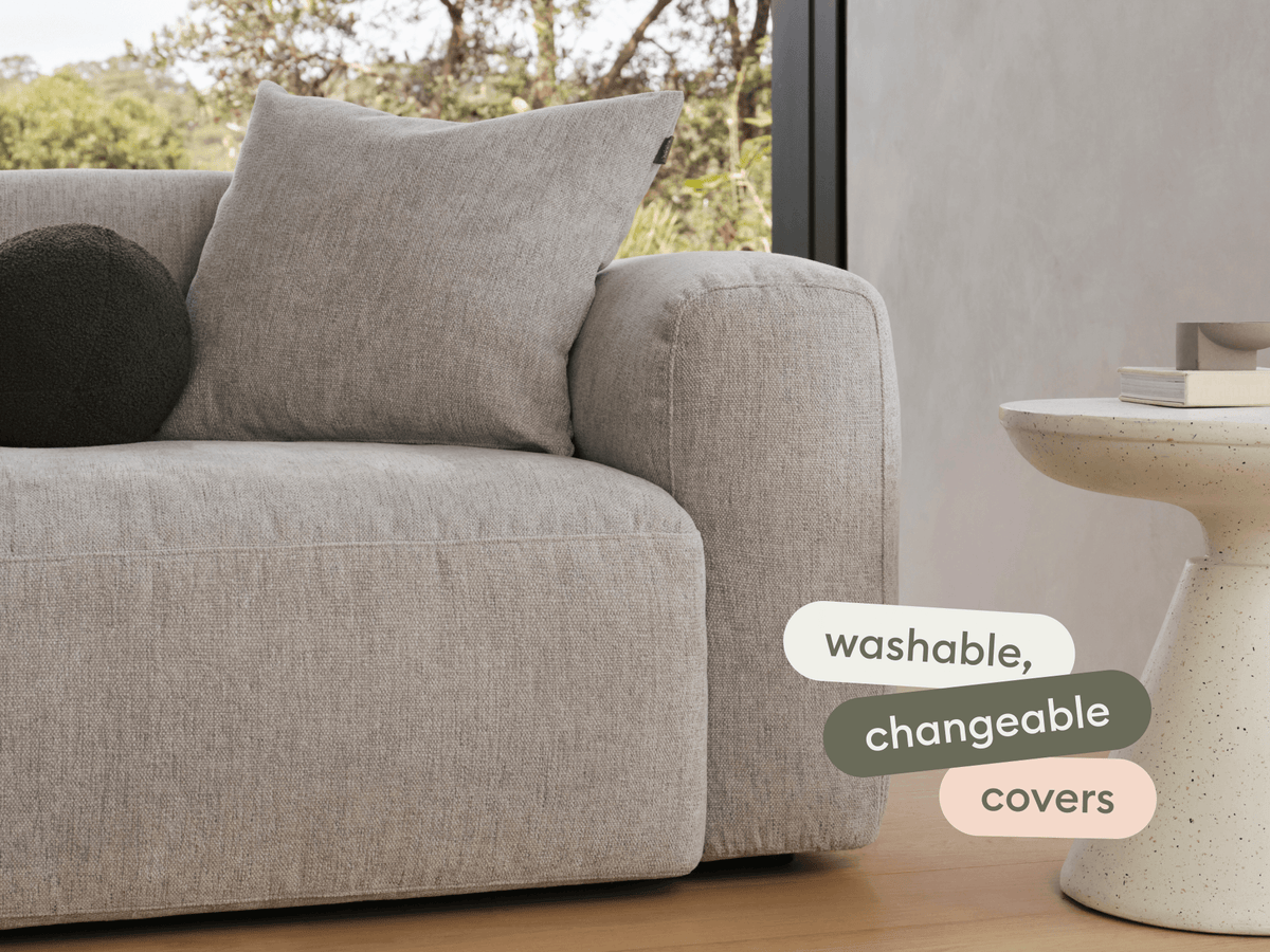 Plush grey fabric sofa with washable, changeable covers, featuring a rounded armrest and cushion. Paired with a minimalist speckled stone side table.