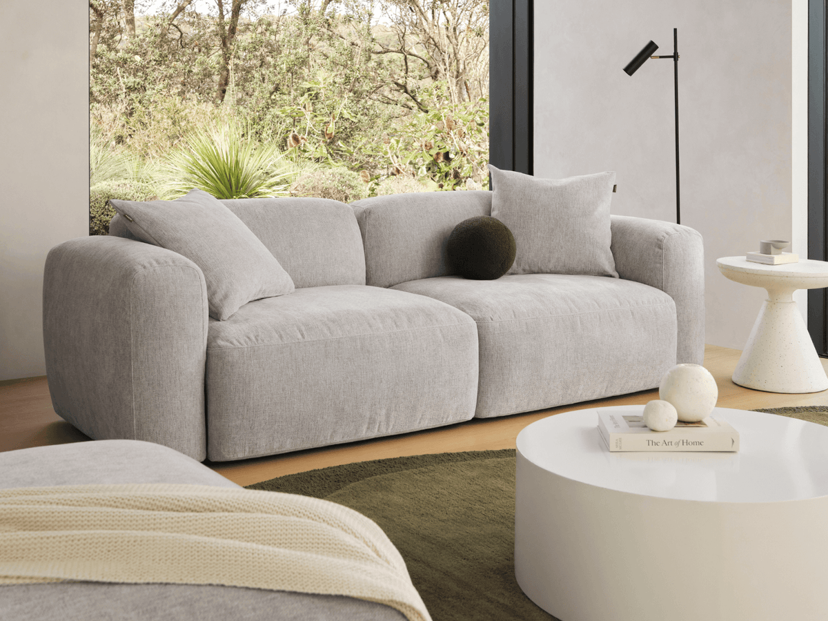 Luxurious grey fabric sofa with plush cushions, curved armrests, and minimalist design. Positioned in a modern living room with natural light and neutral decor.