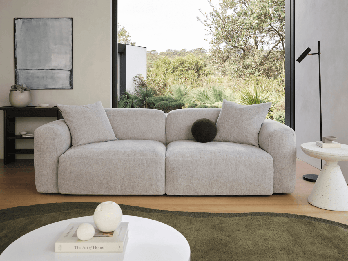 Luxurious modern sofa in soft grey fabric, featuring broad armrests and deep plush cushions. Set in a stylish living room with natural light and minimalist decor.