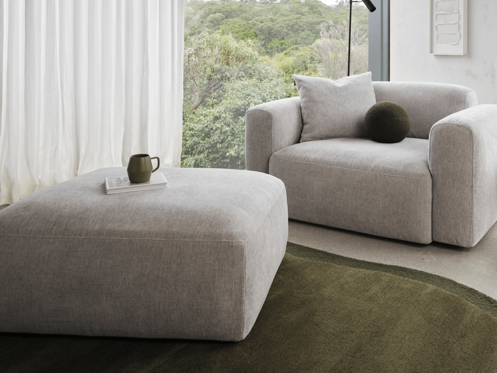 Luxurious grey fabric armchair and ottoman with plush cushions, set on polished concrete flooring against a backdrop of lush greenery and sheer curtains.
