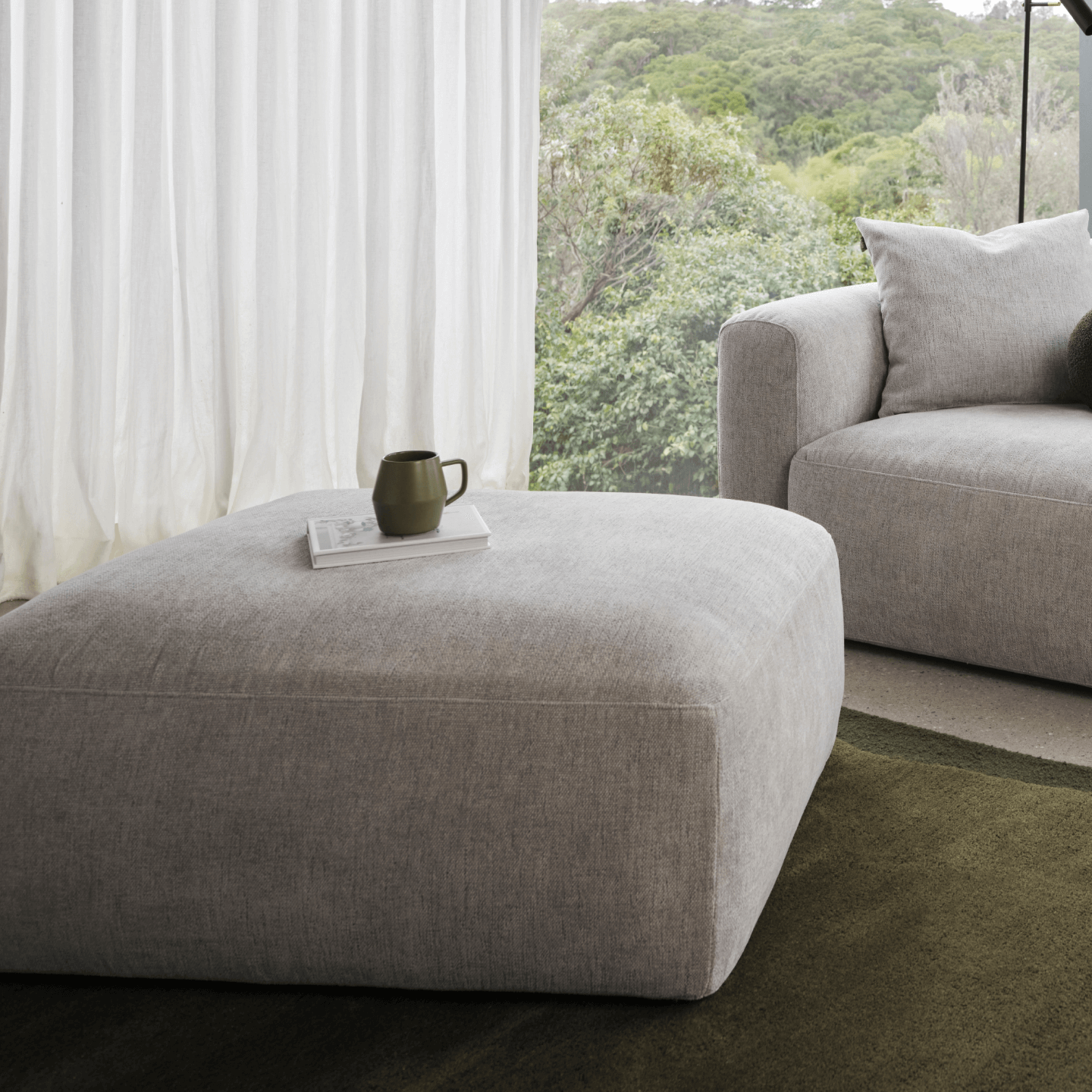 Luxurious grey linen ottoman with clean lines, paired with matching sofa. Set in a bright, airy living room overlooking lush greenery.