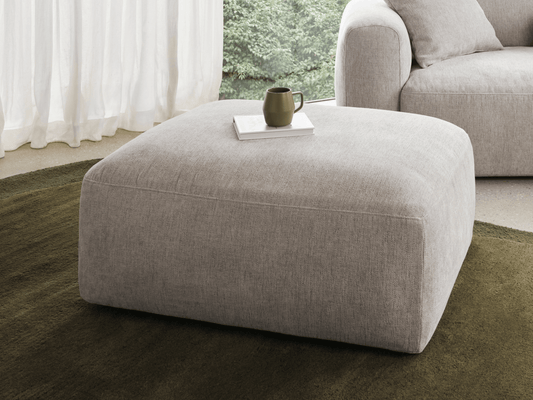 Plush ottoman in light grey fabric, set on a green rug. Features a spacious, cushioned top. Complements modern lounge with floor-to-ceiling windows.