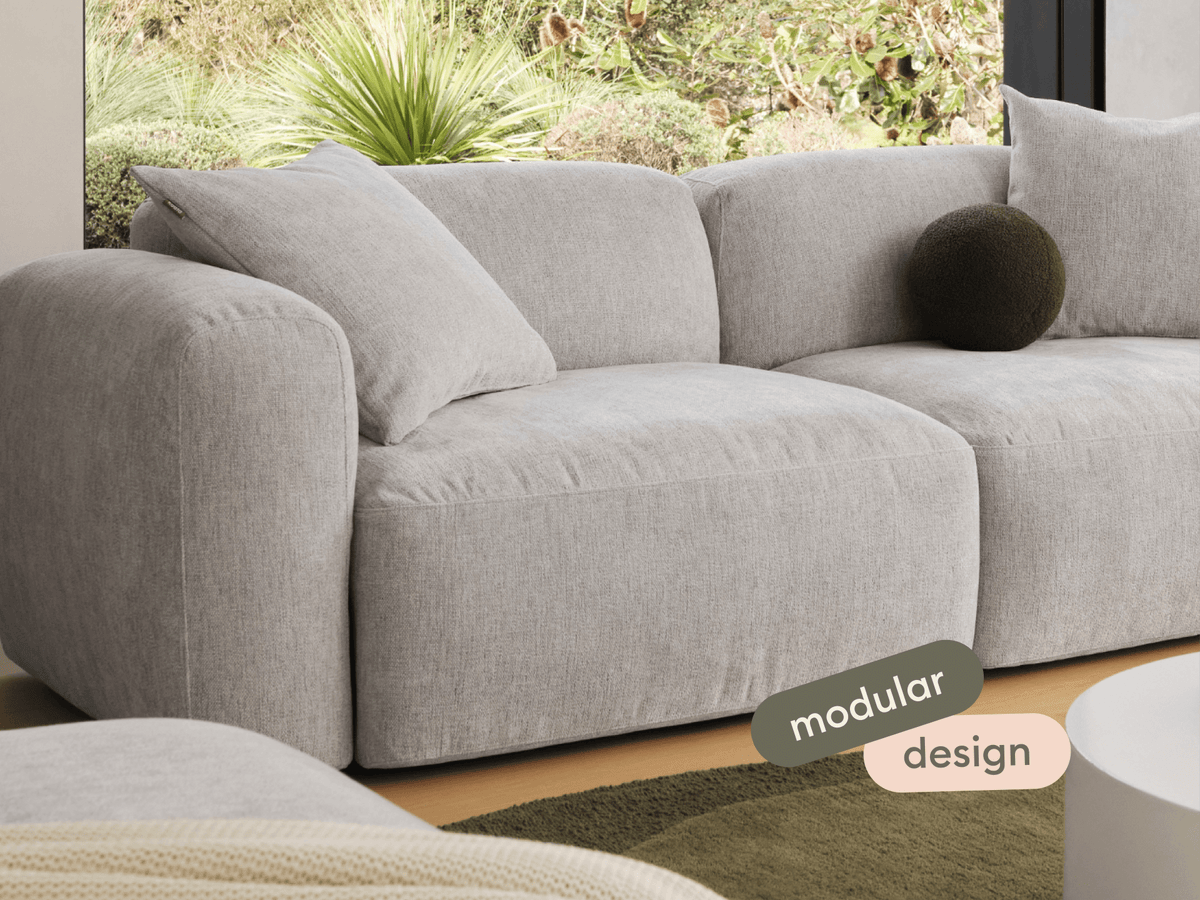 Luxurious modular sofa in soft grey fabric, featuring plush cushioning and rounded armrests. Set in a chic living room with garden views.