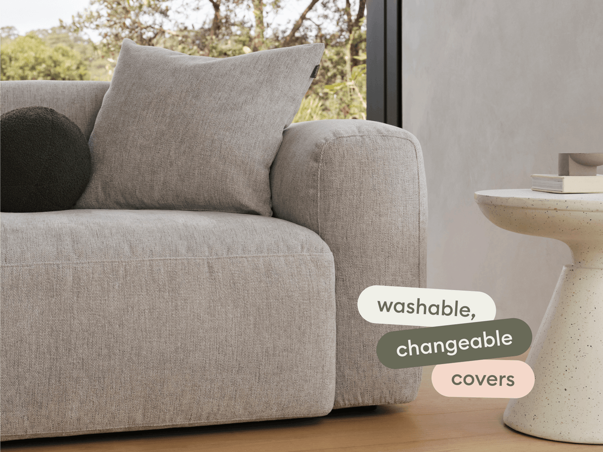 Modern grey linen sofa with plush cushions, complemented by a sleek round stone side table. Set in a bright living space with large windows.