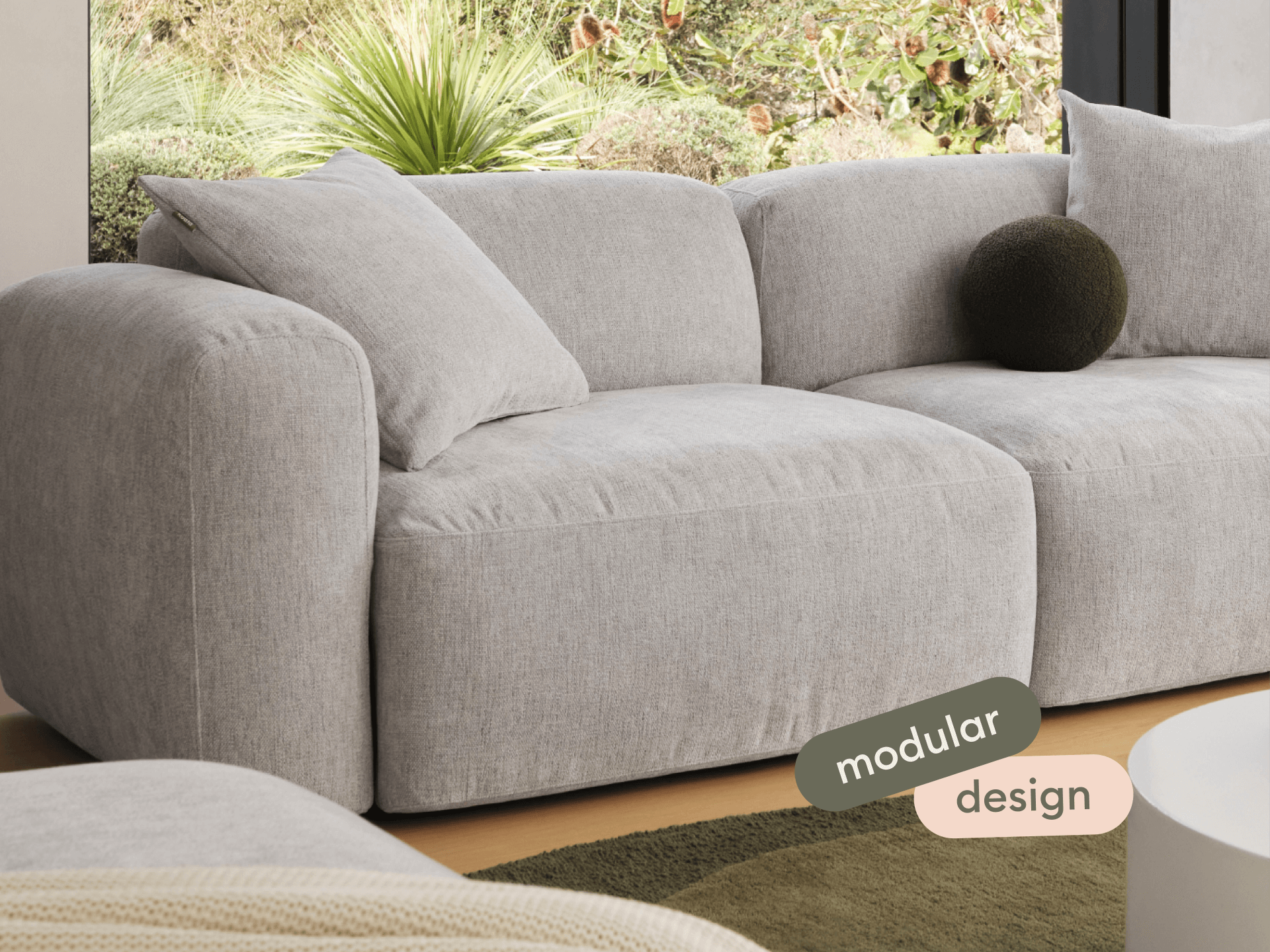 Luxurious modular sofa in soft grey linen, featuring plush cushions and rounded arms. Set in a serene living room with large windows and garden views.