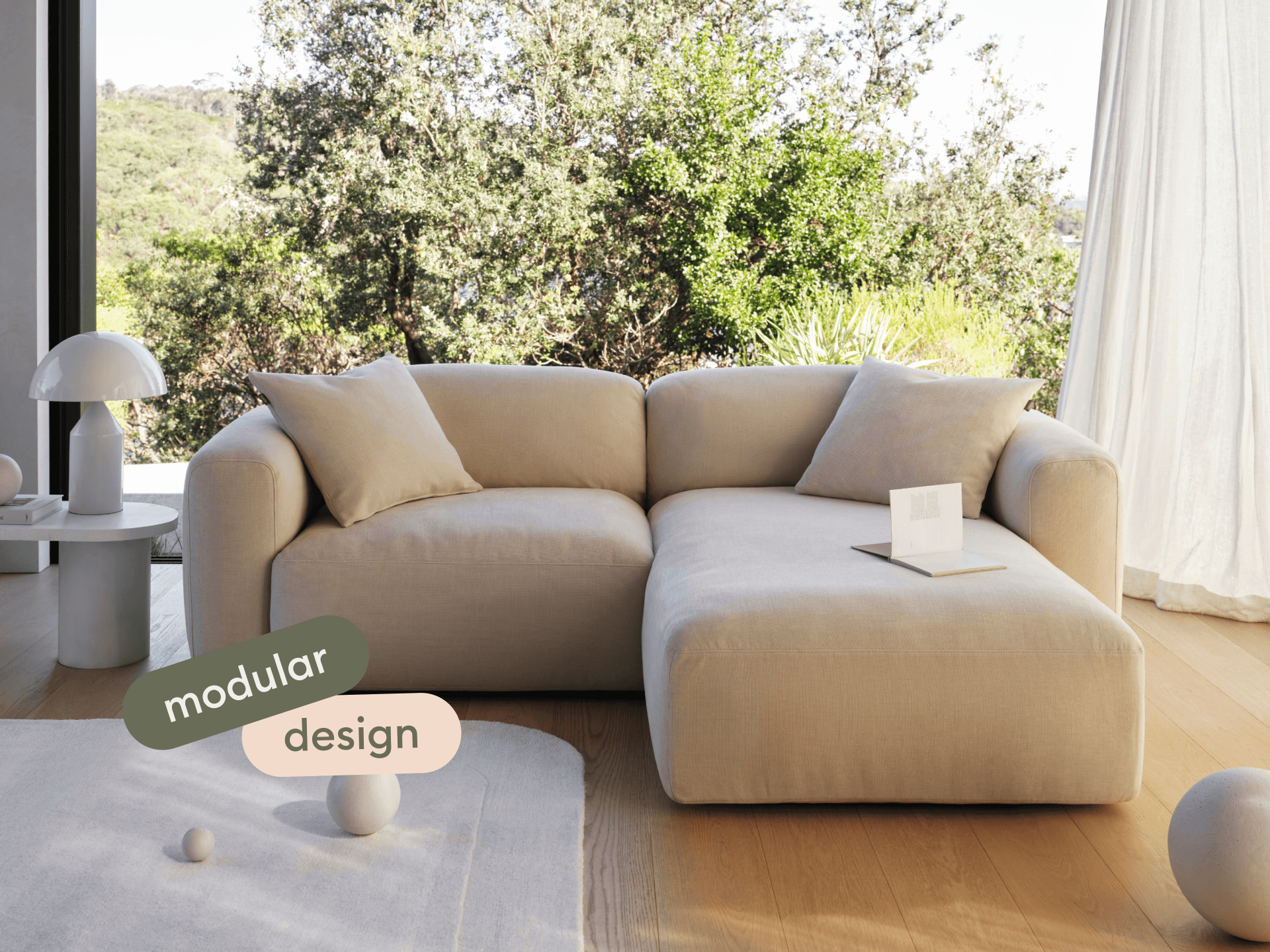 Modular beige fabric sofa with plush cushioning, set on light oak flooring. Minimalist aesthetic, complemented by natural light and serene outdoor view.
