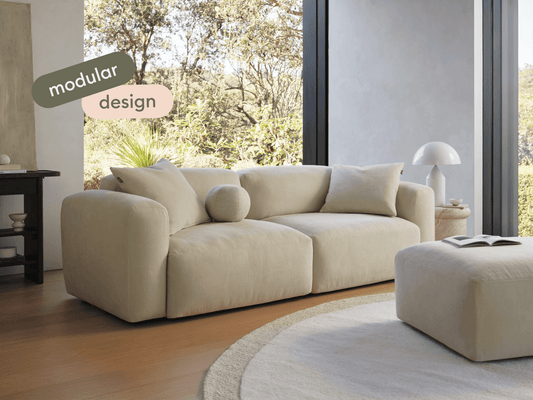 Luxurious modular sofa in cream suede, featuring plush cushions and rounded arms. Set on light wood flooring by large windows, complemented by a matching ottoman.