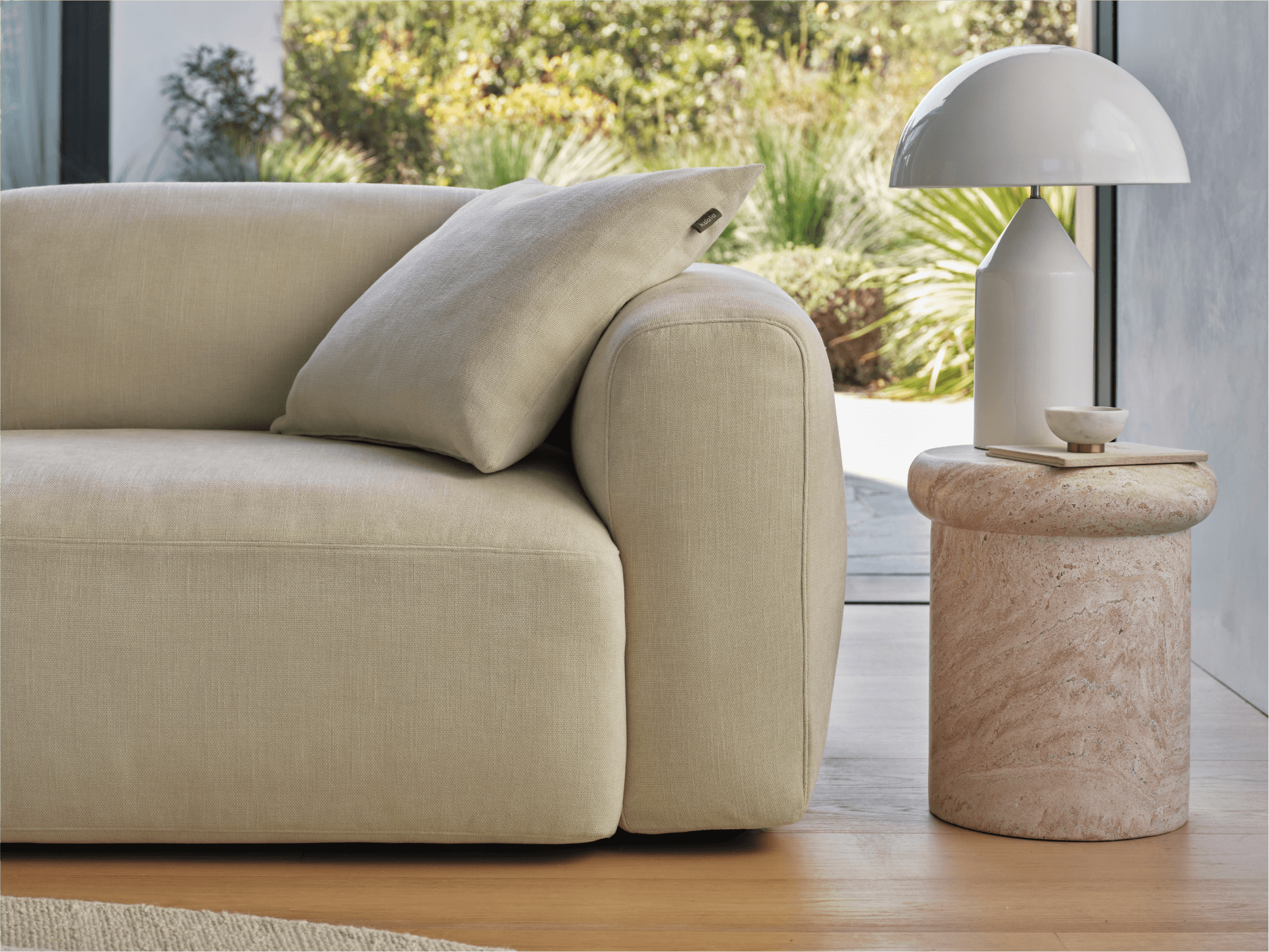 Beige linen sofa with plush cushions; features rolled arms for comfort. Set on light timber flooring beside a round travertine side table and white lamp.