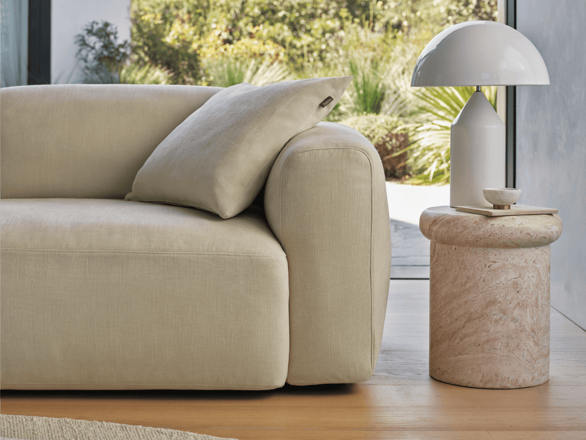 Luxurious beige linen sofa with plush cushions, paired with a cylindrical travertine side table. Set in a sunlit living room with garden views.