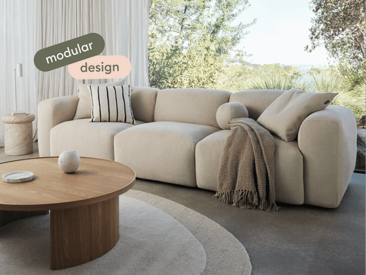Beige modular sofa in premium fabric with plush cushions, set in a light-filled space. Paired with a round wooden coffee table and soft neutral decor.