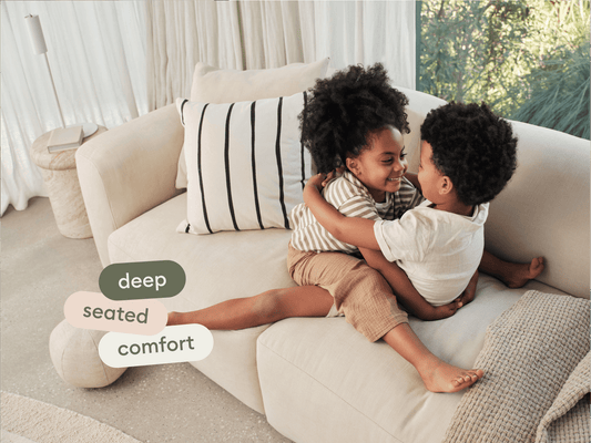 Modern beige sectional sofa with plush fabric upholstery, deep seating, and minimalist design. Set in a light, airy living room with sheer curtains.